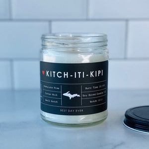 May include: A clear glass jar candle with a black label that says "KITCH-ITI-KIPI" and features a small outline of Michigan's Upper Peninsula. The label also includes details like "Phthalate Free", "Cotton Wick", "Small Batch", "Burn Time 50 hrs", "Soy Based Candle 7 oz", "Batch 1813", and "BEST DAY EVER".