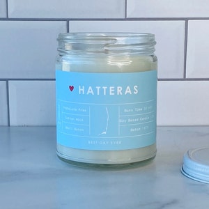 Op de afbeelding: Een blauwe en witte kaars pot met de tekst "HATTERAS" en "BEST DAY EVER". De kaars is op basis van soja en heeft een katoenen lont. De pot heeft een wit deksel.