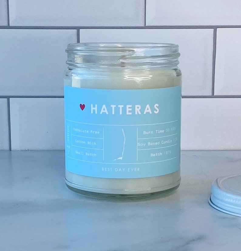 Op de afbeelding: Een blauwe en witte kaars pot met de tekst "HATTERAS" en "BEST DAY EVER". De kaars is op basis van soja en heeft een katoenen lont. De pot heeft een wit deksel.