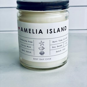 May include: A white candle jar with a black lid and a white label that says "Amelia Island" and has details about the candle, including "Phthalate Free", "Cotton Wick", "Small Batch", "Burn Time 50 HRS", "Soy Based", "Batch 1562", and "Best Day Ever".