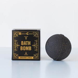 May include: A black bath bomb and a black box with gold lettering. The box reads "BATH BOMB" and "Sandalwood & Vanilla". The bath bomb is round and has a textured surface. The box has an ornate design.