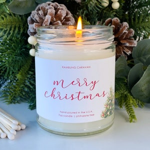 May include: A white candle with a red "Merry Christmas" label and a small green Christmas tree illustration. The candle is in a clear glass jar with a white lid. The label also says "Rambling Caravan", "hand poured in the U.S.A.", "7oz candle", and "phthalate free".