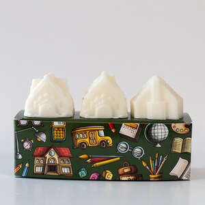 Puede incluir: Tres fundentes de cera blancos con forma de casas en una caja verde con un diseño de temática escolar. La caja está cubierta de ilustraciones coloridas de útiles escolares, incluyendo lápices, un globo terráqueo, un autobús escolar y una calculadora.
