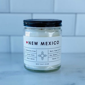 May include: A white candle jar with a black lid and a white label that says "NEW MEXICO" with a red heart. The label also includes details about the candle, such as "Phthalate Free", "Cotton Wick", "Small Batch", "Burn Time 50 Hours", "Soy Based", and "Batch 1912". The label also has a graphic of the New Mexico state symbol. The bottom of the label says "BEST DAY EVER".
