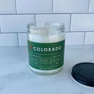 Könnte beinhalten: Ein grünes Glas-Kerzengefäß mit einem weißen Deckel. Das Etikett zeigt "COLORADO" mit einem roten Herzen und einem kleinen Bild der Flagge des Bundesstaates Colorado. Das Etikett zeigt auch "Phthalatfrei", "Baumwolldocht", "Kleine Charge", "Brenndauer 50 Stunden", "Sojabasis" und "Charge 1871". Das Etikett zeigt auch "BEST DAY EVER".