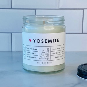 May include: A white candle jar with a black and white label that says "Yosemite" with a red heart. The label also says "Phthalate Free", "Cotton Wick", "Small Batch", "Burn Time 50 HRS", "Soy Based 7 OZ", and "Batch 1864". The label also has a black and white illustration of a mountain.