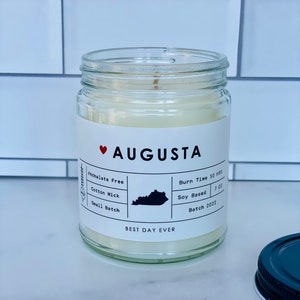 May include: A white candle in a clear glass jar with a black and white label that says "AUGUSTA" with a red heart. The label also says "Phthalate Free", "Cotton Wick", "Small Batch", "Burn Time 50 HRS", "Soy Based", "7 OZ", and "Batch 2022". The label also has a black outline of the state of Kentucky. The bottom of the label says "BEST DAY EVER".