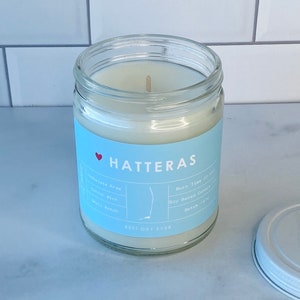 Op de afbeelding: Een witte kaars pot met een blauw label waarop "HATTERAS" en "BEST DAY EVER" staat. Het label bevat ook details over de kaars, zoals "Ftalatenvrij", "Katoenen lont", "Soja-kaars" en "Brandtijd 30 uur".