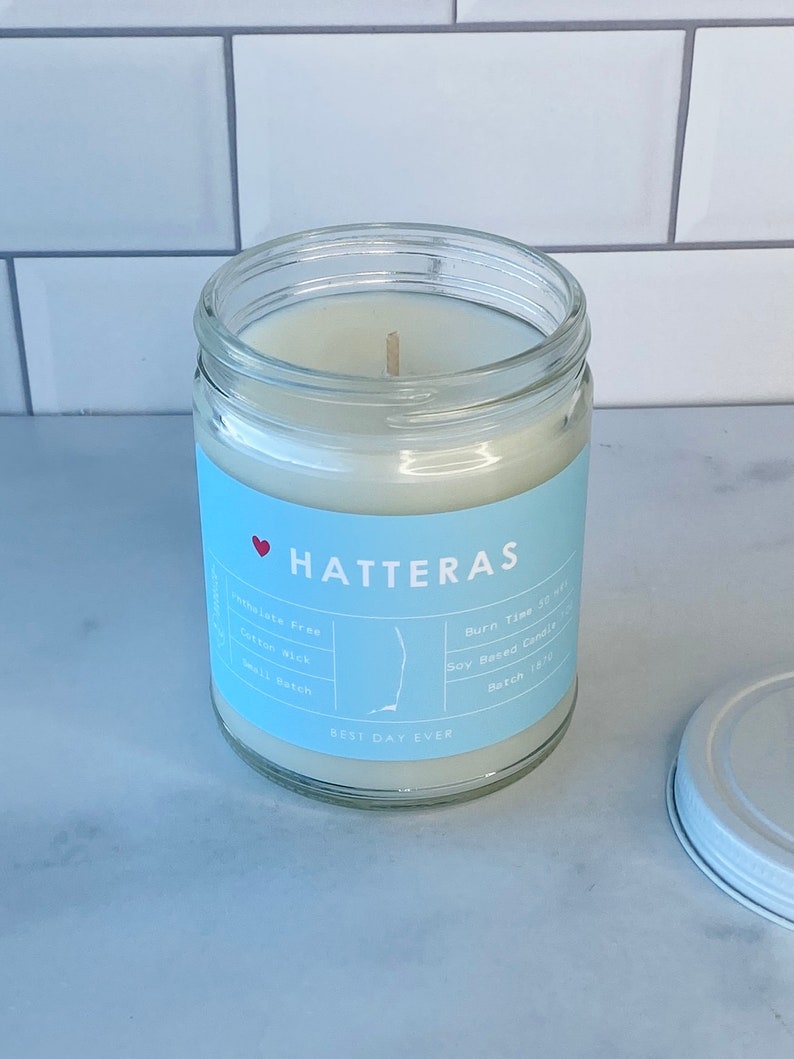 Op de afbeelding: Een witte kaars pot met een blauw label waarop "HATTERAS" en "BEST DAY EVER" staat. Het label bevat ook details over de kaars, zoals "Ftalatenvrij", "Katoenen lont", "Soja-kaars" en "Brandtijd 30 uur".