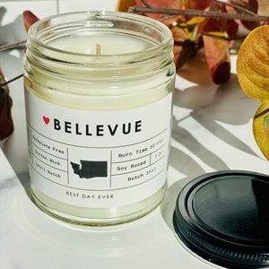 May include: A white candle in a clear glass jar with a black lid. The label says "BELLEVUE" with a red heart and "BEST DAY EVER". The candle is soy-based and has a burn time of 50 hours. The label also says "Small Batch" and "Batch 1953".