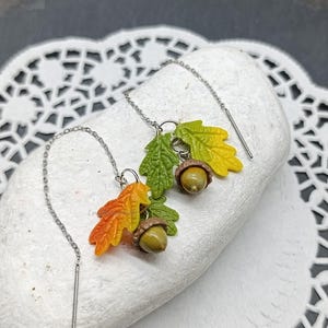Könnte beinhalten: Ohrringe mit Miniatur-Herbst-Anhängern. Jeder Ohrring hat eine silberne Kette mit einem kleinen, baumelnden Eichenblatt- und Eichel-Design. Die Blätter sind in Grün-, Gelb- und Orangetönen bemalt, und die Eicheln sind braun.