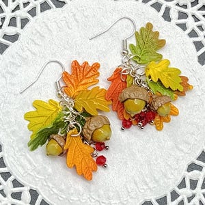 Może przedstawiać: Para kolczyków o tematyce jesiennej z kolorowymi liśćmi w odcieniach pomarańczu, żółci i zieleni, a także brązowymi żołędziami i małymi czerwonymi koralikami. Kolczyki mają srebrne haczyki.