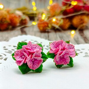 Peut inclure: Une paire de boucles d'oreilles artisanales en forme de fleurs roses, avec des pétales détaillés et des feuilles vertes. Les boucles d'oreilles sont présentées sur un fond flou, suggérant un thème floral. Un accessoire unique.