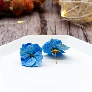 Peut inclure: Une paire de boucles d'oreilles clous en forme de fleurs d'hortensia bleu clair. Les boucles d'oreilles présentent des pétales détaillés et une petite tige dorée. Elles sont présentées sur une surface blanche.