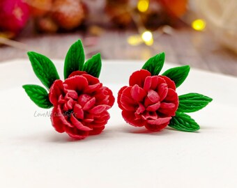 May include: A pair of red flower earrings with green leaves. The earrings are in the shape of a peony flower. The earrings are on a white surface. The text "LoveNatureGifts" is visible on the left earring.