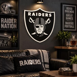 May include: A large wall-mounted Raiders shield sign featuring the team logo in black, white, and grey. The shield displays the word RAIDERS at the top above a central illustration of a football player wearing a helmet and an eye patch.