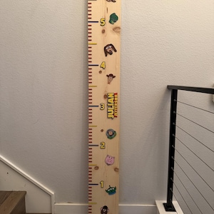 May include: A vertical wooden growth chart ruler featuring a natural light wood grain finish. The board displays printed numbers from 1 to 6 in yellow, with red and yellow tick marks. It includes colorful character icons and the name JULIAN JEFFERSON.