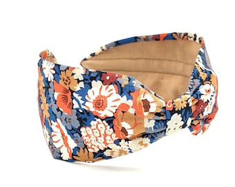 May include: A floral print headband with a knotted design. The headband features a pattern of white, orange, and brown flowers on a navy blue background. The interior is a light brown fabric.