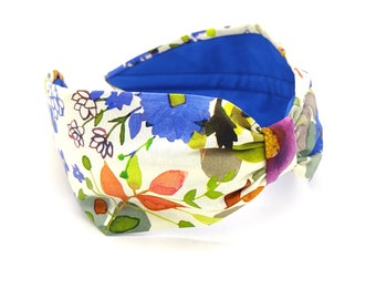 May include: A white headband with a floral print in shades of blue, green, orange, and pink. The headband has a knotted detail and a blue lining.
