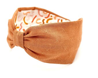 May include: A brown fabric headband with a knotted design. The headband has a subtle pattern of small crosses.