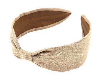 May include: A tan fabric headband with a knotted design. The headband has a wide band and a brown interior. The fabric has a subtle cross-stitch pattern. The headband is designed to be worn around the head.