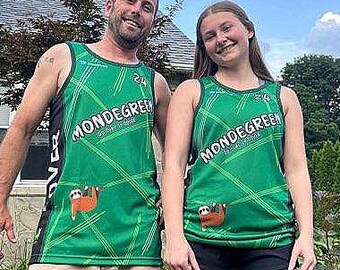 Puede incluir: Dos personas con camisetas sin mangas verdes a juego con el texto "MONDEGREEN" y un gráfico de perezoso. Las camisetas sin mangas tienen un diseño geométrico. Una persona lleva pantalones cortos color caqui, la otra pantalones cortos negros. El fondo incluye una casa, árboles y un cielo azul.