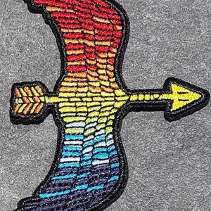 May include: A rainbow-colored embroidered patch of a bird with an arrow through it. The bird's wings are spread out and the arrow is pointing to the right. The patch is on a gray background.