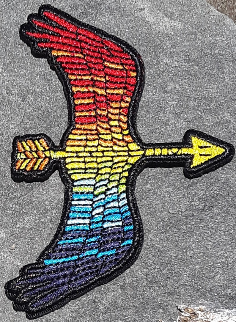 Op de afbeelding: Een geborduurde patch met een regenboogkleurige vogel met een pijl erdoorheen. De vleugels van de vogel zijn uitgespreid en de pijl wijst naar rechts. De patch is op een grijze achtergrond.