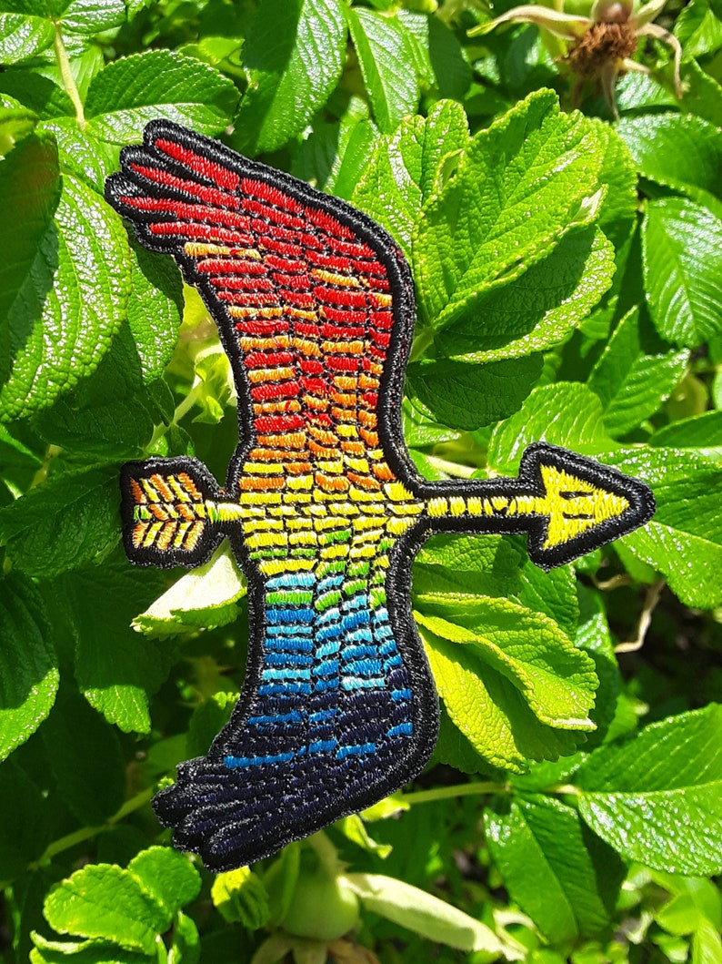 Op de afbeelding: Regenboogkleurige geborduurde strijkpatch van een vliegende vogel met een pijl in zijn bek.