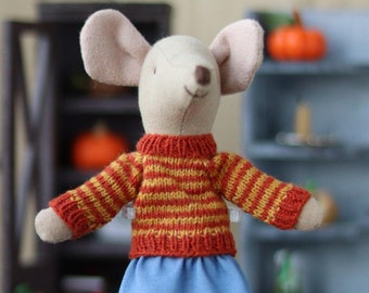 May include: A small, plush mouse doll wearing a red and yellow striped sweater, a blue skirt, and brown shoes. The mouse is standing on a clear display stand and there is a small orange pumpkin in the foreground.