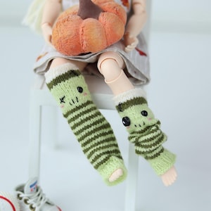 May include: A doll with a pumpkin and green striped leg warmers with faces. The doll is wearing a dress and white trainers with red trim. The pumpkin is orange and brown.