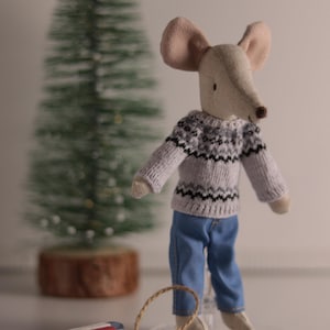 May include: A stuffed mouse doll wearing a gray sweater with a black and white pattern and blue jeans, standing next to a small wooden sled with red and white stripes. A small Christmas tree is in the background.