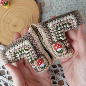 May include: A small crocheted cardigan with a brown, green, and white color scheme. The cardigan has two embroidered mushroom designs with red tops and white spots.