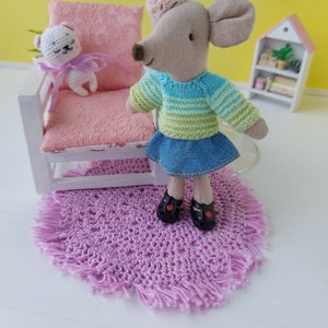 May include: A plush gray mouse doll wearing a blue and green striped sweater and a denim skirt. The doll is standing on a pink crocheted rug with fringe. A small white plush bear doll is sitting on a pink chair in the background.