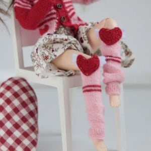 Pode incluir: Uma boneca com cabelo castanho em rabos de cavalo, vestindo um cardigã listrado vermelho e rosa, uma saia floral e polainas rosa com corações vermelhos. Um enfeite em forma de coração xadrez vermelho e branco está por perto. A boneca está sentada em uma cadeira branca.