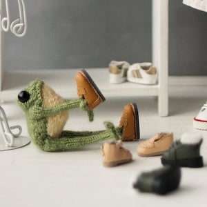 May include: A doll with brown hair and a white dress with butterfly designs is standing in front of a white clothing rack. The doll is wearing orange and brown striped tights and white sneakers. There are several miniature shoes and a knitted frog on the floor.
