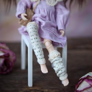 Può includere: Una bambola che indossa un vestito lavanda con pizzo bianco e scaldagambe bianchi e verdi. La bambola è seduta su uno sgabello bianco.