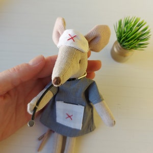 Op de afbeelding: Een pluche muis speelgoedje verkleed als dokter, met een witte hoed met een rood kruis, een grijs doktersuniform met een witte zak en een stethoscoop.