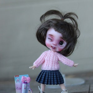 May include: A doll with brown hair wearing a pink sweater and a blue plaid skirt. The doll is winking and smiling. The doll is holding a pink and white microphone with the text "Jojo Siwa Light Up Microphone" on the packaging.