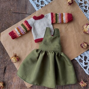 May include: A green corduroy dress with a ruffled skirt and a cream colored sweater with colorful stripes on the sleeves.