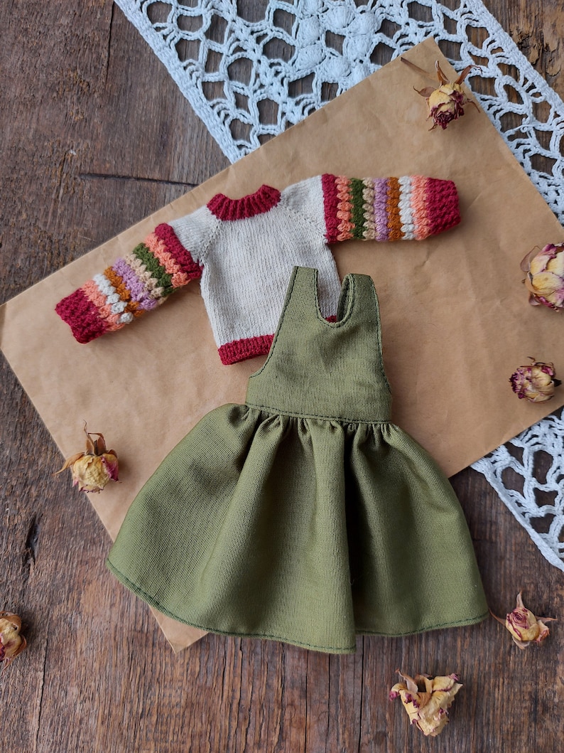 May include: A green corduroy dress with a ruffled skirt and a cream colored sweater with colorful stripes on the sleeves.