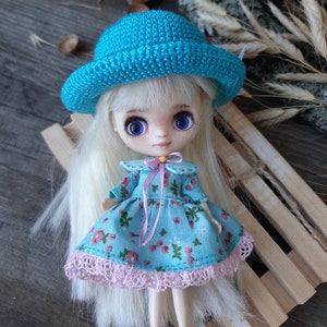 Pu&ograve; includere: Una piccola bambola con lunghi capelli biondi che indossa un cappello all'uncinetto blu, un vestito floreale blu e bianco con un bordo in pizzo rosa e un nastro rosa intorno al collo. La bambola &egrave; seduta su una superficie di legno.