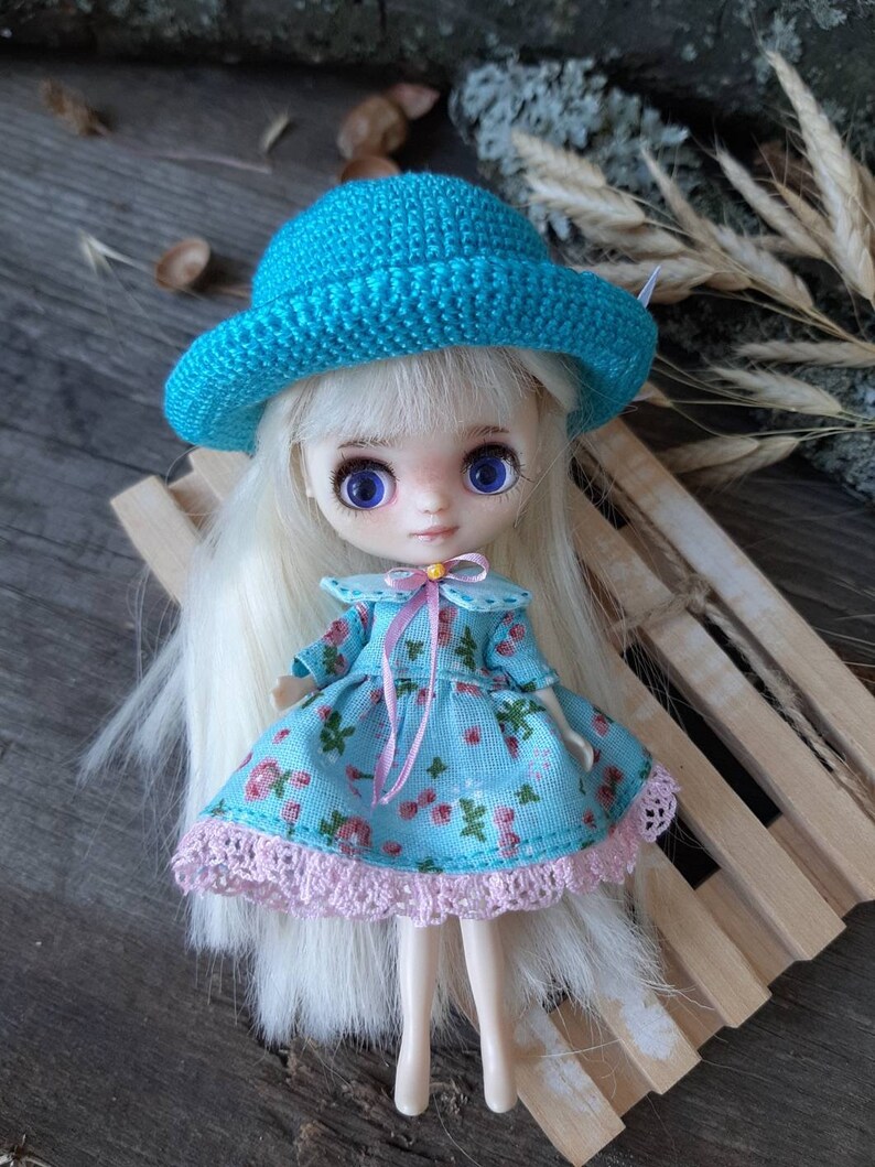 Pu&ograve; includere: Una piccola bambola con lunghi capelli biondi che indossa un cappello all'uncinetto blu, un vestito floreale blu e bianco con un bordo in pizzo rosa e un nastro rosa intorno al collo. La bambola &egrave; seduta su una superficie di legno.
