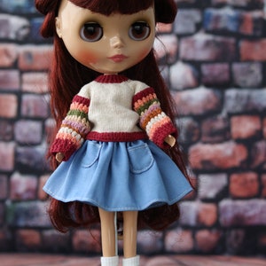 May include: A doll with long, dark red hair wearing a white sweater with colorful sleeves, a blue denim skirt, and brown shoes. The doll is standing on a green stand.