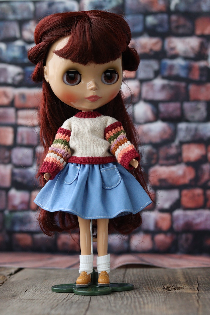 May include: A doll with long, dark red hair wearing a white sweater with colorful sleeves, a blue denim skirt, and brown shoes. The doll is standing on a green stand.