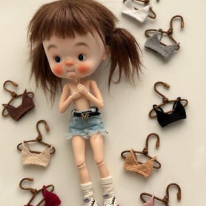 Può includere: Una bambola con capelli castani a codini, che indossa pantaloncini di jeans, calzini bianchi e scarpe da ginnastica. Intorno alla bambola ci sono diversi reggiseni in miniatura lavorati a maglia in vari colori, ciascuno appeso a una piccola gruccia di legno.