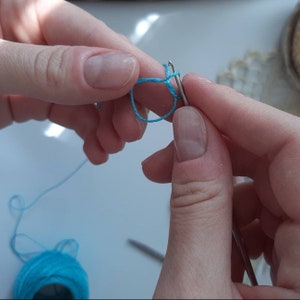 Pu&ograve; includere: Primo piano di una mano che tiene un uncinetto e filo blu, creando un nodo scorso. Il filo &egrave; avvolto attorno all'uncinetto e l'estremit&agrave; del filo &egrave; tenuta nell'altra mano.
