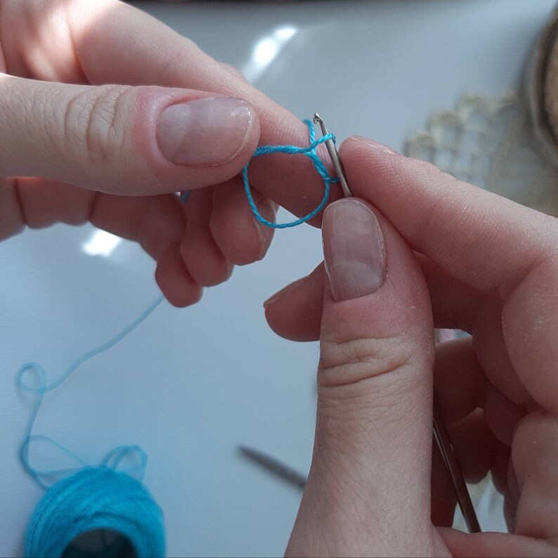 Pu&ograve; includere: Primo piano di una mano che tiene un uncinetto e filo blu, creando un nodo scorso. Il filo &egrave; avvolto attorno all'uncinetto e l'estremit&agrave; del filo &egrave; tenuta nell'altra mano.