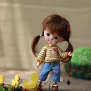 May include: A small doll with brown hair in pigtails wearing a yellow and white striped sweater and blue jeans pulls a toy train of yellow ducks on wheels.