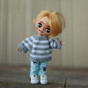 May include: A small doll with blonde hair wearing a gray and white striped sweater, blue pants with black stars, and white sneakers.